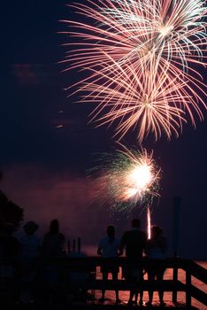 Photograph of various firework bursts from a 4th of July Celebration on Saint Simons Island Georgia.  Fireworks are shot off over water and reflection backlights people in foreground.