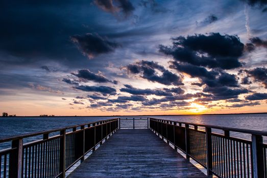 Photograph of a backlit pier near Buffalo New York taken at sunset with multilayered clouds and sun visible.