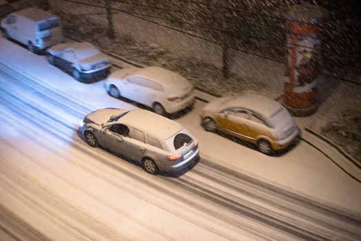 View from above on cars parked along a street and one car driving at night in winter snow