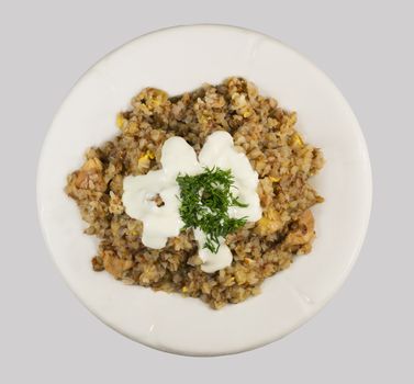 Buckwheat porridge, mixed with grated egg and poured sour cream with dill on white plate. Isolated on a light gray background.
