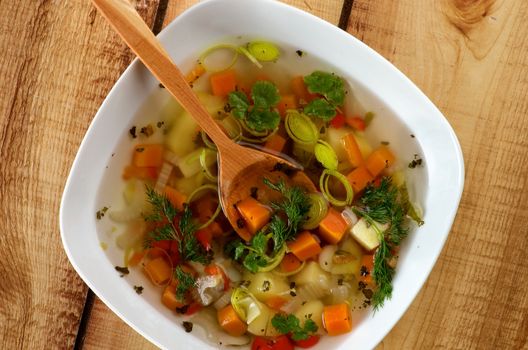 Vegetable Soup with Potato, Carrot, Bell Pepper, Leek, Parsley and Dill with Wooden Spoon in White Bowl closeup on Wooden background. Top View