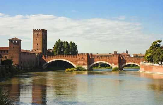 River bank view of the famous The Castelvecchio Bridge in Verona, Italy in a bright sunny day with white clouds in the distance and colorful reflections over the water surface.