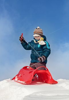 Boy preparing for a ride with red plastic sleigh on a snowy hill in sunny winter day