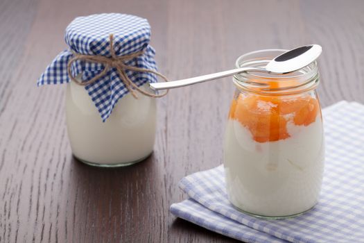 Homemade milk yogurt with peach or apricot jam in glass pot and metal spoon served on linen napkin, wooden table