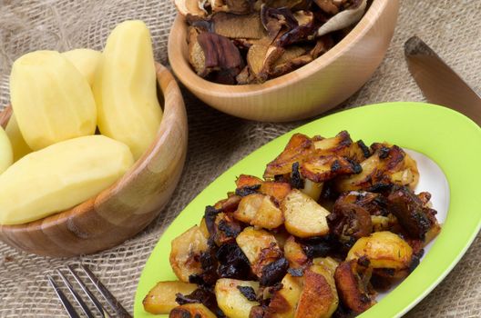Roasted Potato with Mushrooms, Onion and Spices. Arrangement of Prepared Potato on Green Plate and Two Wood Bowls with Raw Potato and Slices of Mushrooms and Fork with Knife close up