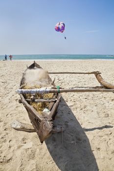 Horizontal landscape of a Goan beach in India with a moored fishermans boat on a sandy beach, bathers about to undress an get in the ocean while a paraglider drifts by