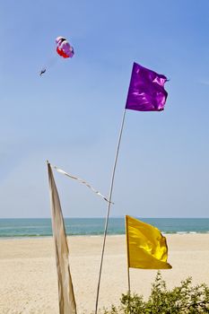 Vertical generic shot of colorful flags blowing in the breeze outside a shack restaurant in Goa India. Sandy beach with surf and coastline, picturesque idyllic scene and a paraglider passes by in the distance