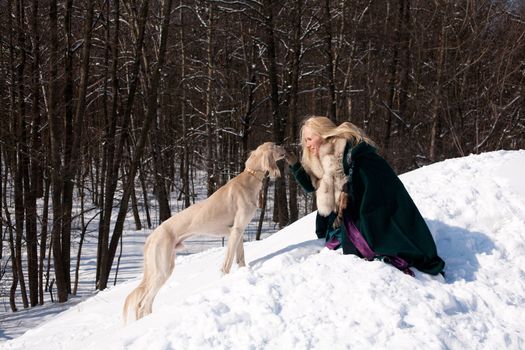 A blonde girl and a standing white saluki on snow
