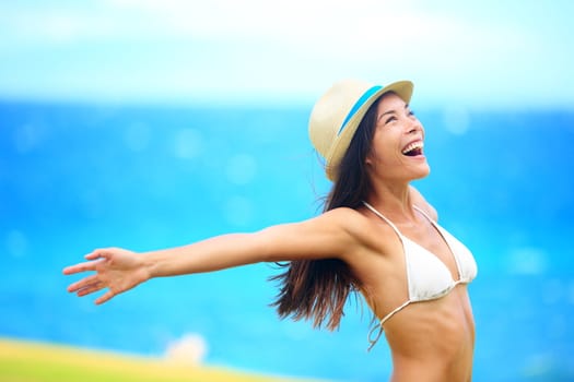 Freedom - free young woman happy on beach smiling joyful and cheerful with arms out on blue ocean sea background. Multicultural woman wearing beach hat and bikini.
