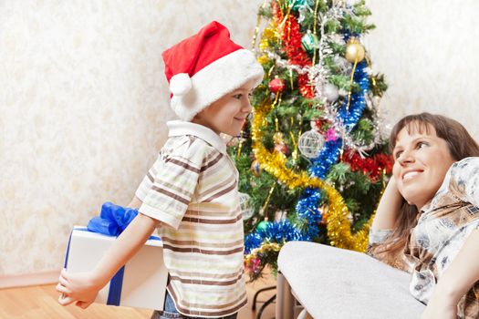 Little son wearing Santa Claus hat giving new year present or christmas holiday gift box to smiling mother 