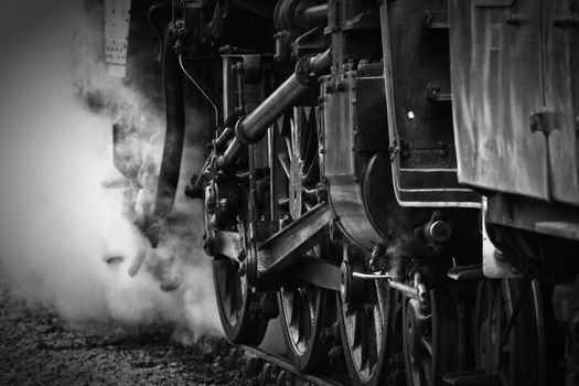 Black and white image of the wheels of an old vintage steam engine or locomotive on the tracks with clouds of steam