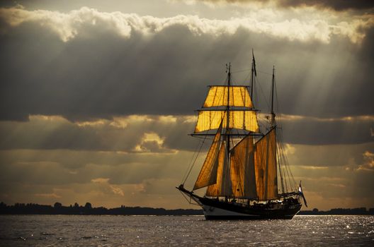 A three-master sailing on the Elbe near Hamburg. The evening light breaks through the gaps in the clouds behind the ship, illuminating the sails from behind. Processed to give it an antique look.