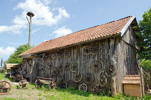 retro carriage wheel hang on rural barn house. wooden benches. stork nest on top.
