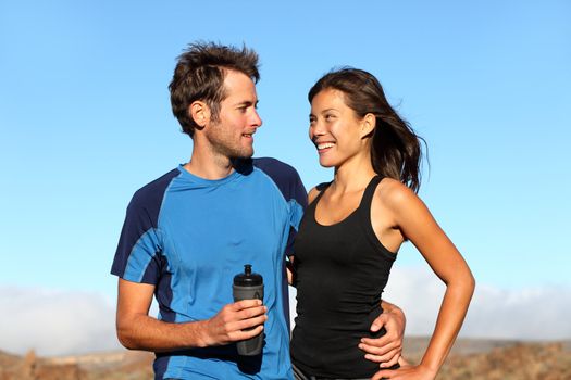 Young romantic healthy athletic couple standing arm in arm taking a break from training in open countryside smiling into each others faces. Multiethnic couple with Asian woman and Caucasian man.