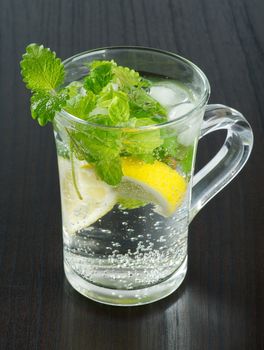 Glass Cup of Beverage with Mint Leafs, Lemon and Ice Cubes closeup on Black Wood background 