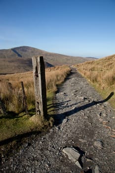 A footpath leads from a post into the distance with a mountain against a blue sky.