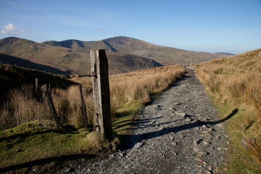 A footpath leads from a post into the distance with a mountain against a blue sky.