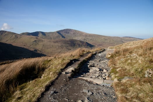 A mountain footpath with a drainage pipe crossing with mountains in the distance.