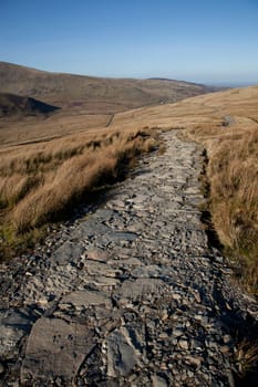 A well maintained stone made footpath with a grassy landscape leading towards hills.