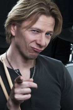 Man in front of a black background an his drums is holding drumsticks and is pointing at viewer