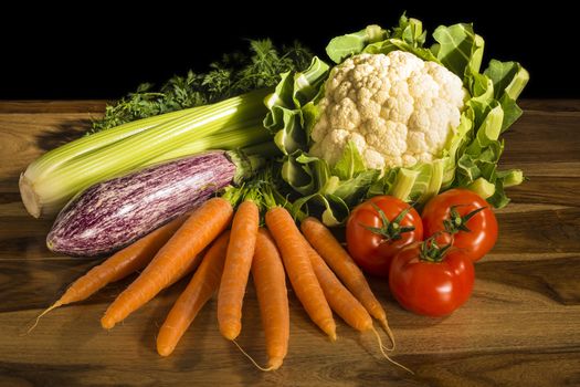 Vegetables for cooking with cauliflower, carrots, tomatoes, celery and eggplant on a wooden table and black background