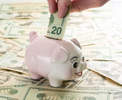 Fingers placing a single $20 bill into the slot on a piggy bank with $20 dollar notes on table
