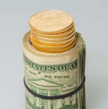 Gold coins standing on roll of many $20 dollar bills and notes secured by elastic band on white table
