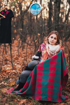 Beautiful girl sitting on armchair in autumn forest with cup in her hands