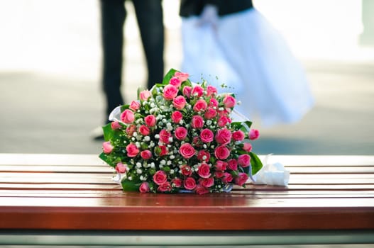 Wedding bouquet from the roses laying on a bench against unrecognizable groom and bride. Blur background. 