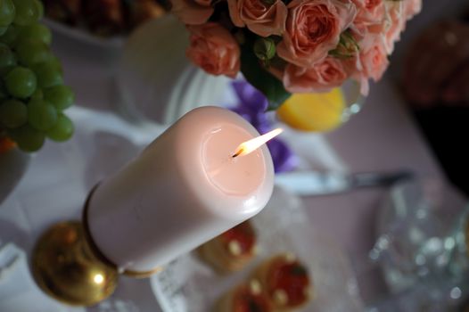 Burning candle standing on table in environment of meal and flowers. Romantic conditions. Holiday decoration