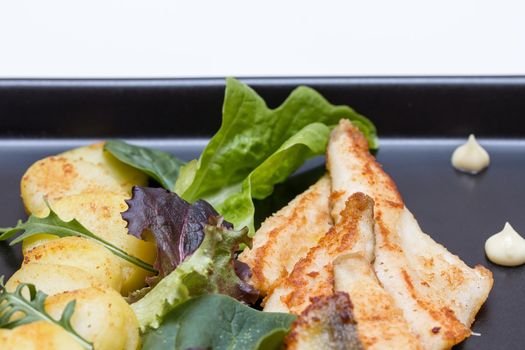 Fried perch filets with potatoes, lemon and salad in a black plate