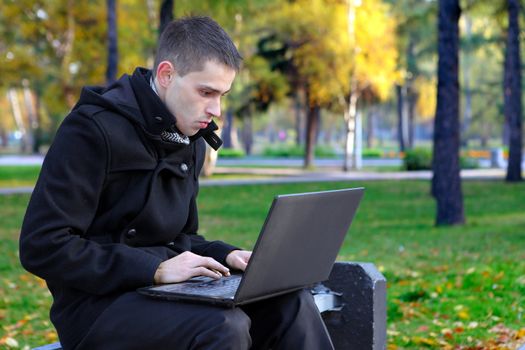 Young man with Laptop sitting on the bench at the Autumn Park