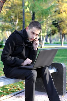 Busy Young man with Laptop sitting on the bench at the Autumn Park and Talking on Mobile Phone