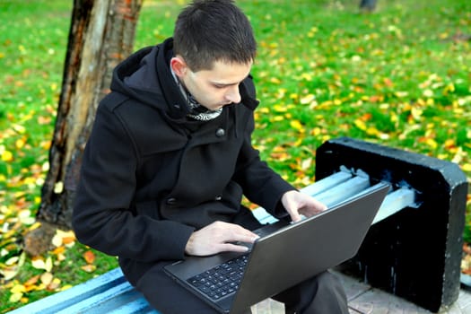 Young man with Laptop sitting on the bench at the Autumn Park