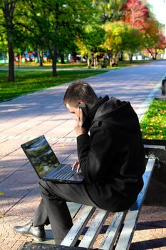Busy Young man with Laptop sitting on the bench at the Autumn Park and Talking on Mobile Phone