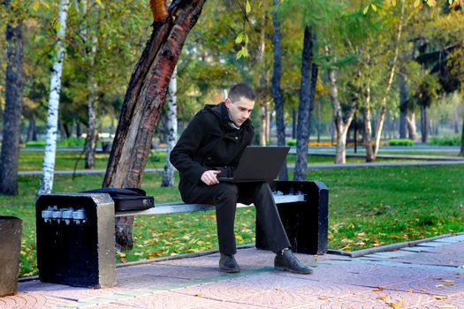 Young man with Laptop sitting on the bench at the Autumn Park