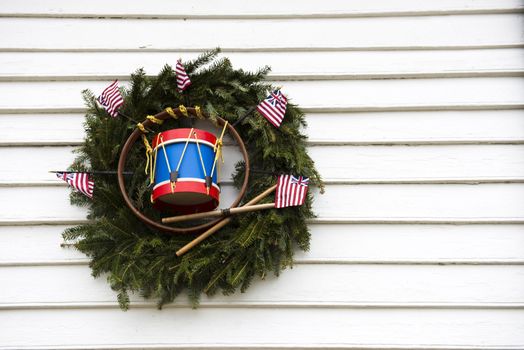 Pine Wreath with toy flags and drum hanging on rustic white wood siding.