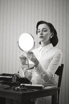 Retro secretary checking her make up on while on her workplace, she is holding a mirror