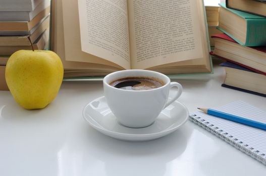 A cup of coffee and an apple on a table of books with a notebook and pencil