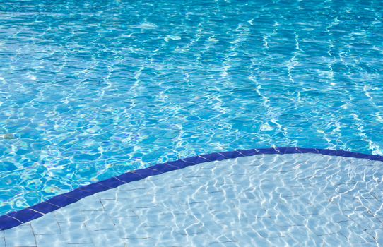 Background of clear blue water with sun reflections in an outdoor swimming pool with two different depths, a shallow and a deep depth.
