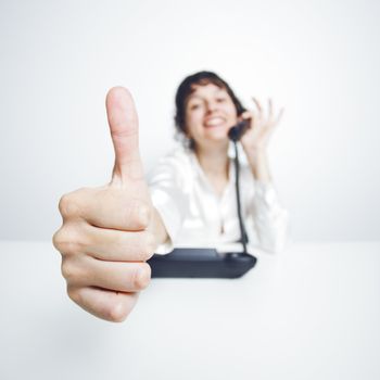 thumbs up shown by a happy, smiling young woman working at her office desk while phoning
