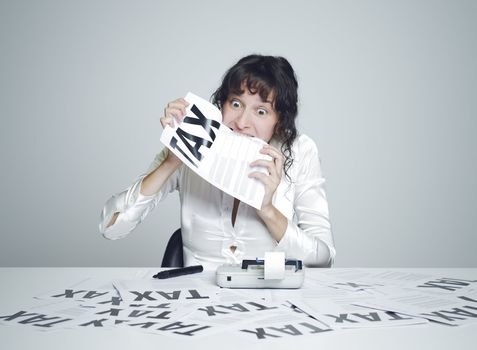 Young desperate woman at her paperwork covered desk biting a tax form