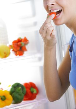 Young woman eating a strawberry near the refrigerator, close up