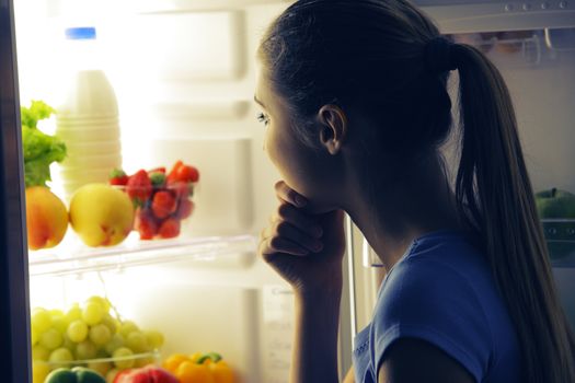 Young woman craving food choosing near refrigerator at night