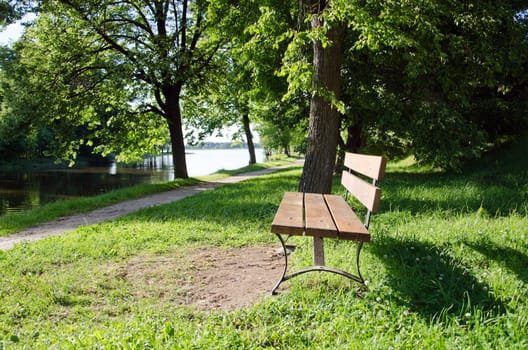 Wooden bench near lake resort. Small path between trees.
