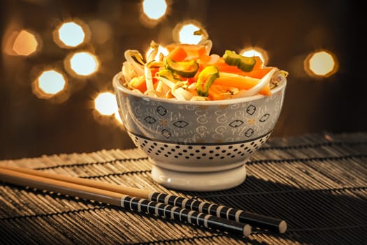 Shell with Asian vegetables and chopsticks on black bamboo mat and lights in the background