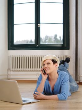 Photo of a beautiful young female lying on the floor working on a laptop and talking on a smartphone.