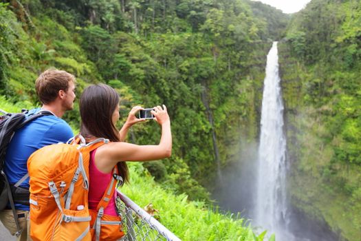 Couple tourists on Hawaii by waterfall. Tourist taking photo pictures of Akaka Falls waterfall on Hawaii, Big Island, USA. Travel tourism concept with multicultural tourist couple.