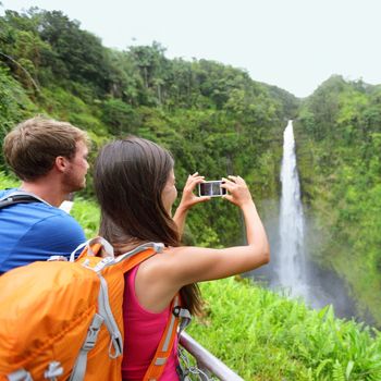 Tourist couple on Hawaii taking pictures of the famous Akaka Falls waterfall on Hawaii, Big Island, USA. Happy cheerful young multicultural couple tourists on travel.