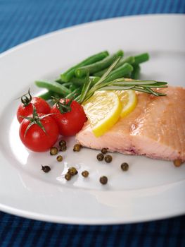 Photo of a cooked salmon steak with rosemary, lemon slices, cherry tomatoes, green beans, and peppercorns on a white plate.